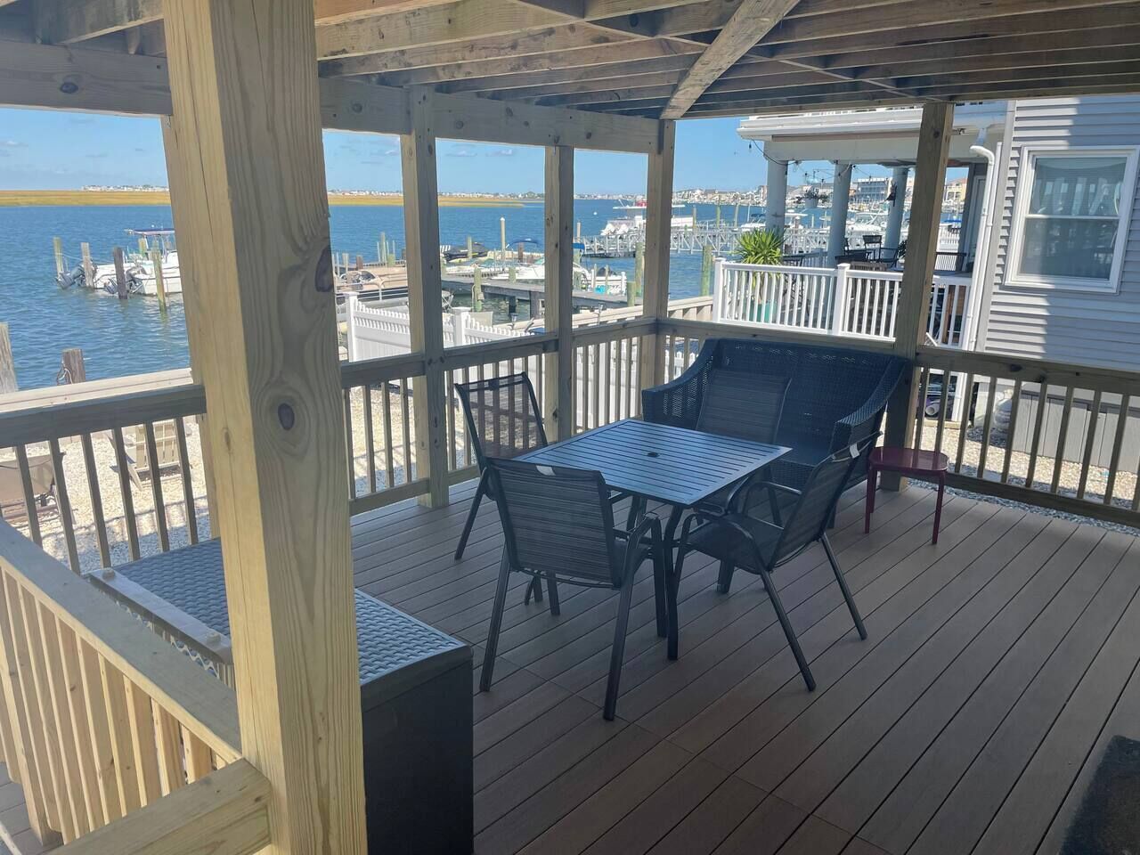 A covered outdoor deck with a table and chairs, overlooking a marina with boats and a distant shoreline.