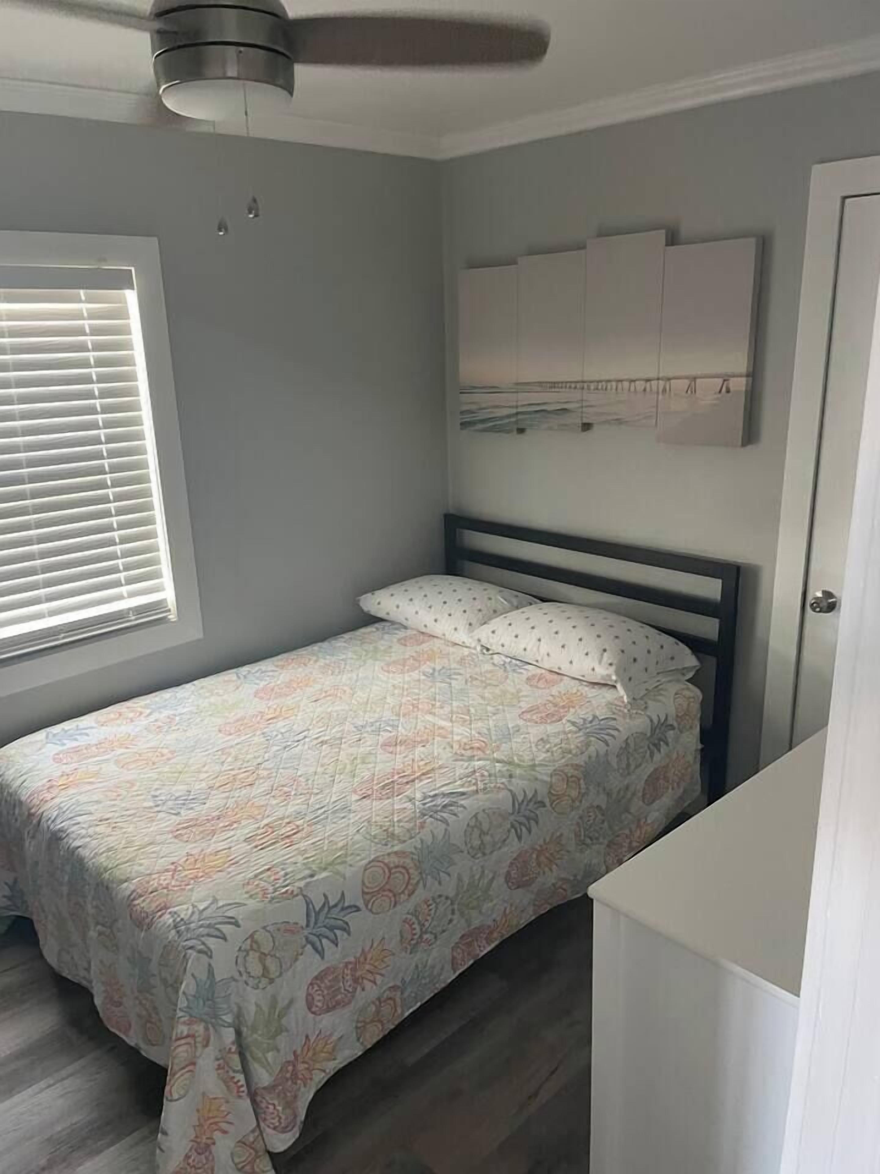 A single bed with a floral bedspread, a ceiling fan, a window with blinds, and a wall-mounted artwork.