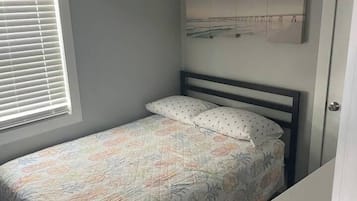 A single bed with a floral bedspread, a ceiling fan, a window with blinds, and a wall-mounted artwork.