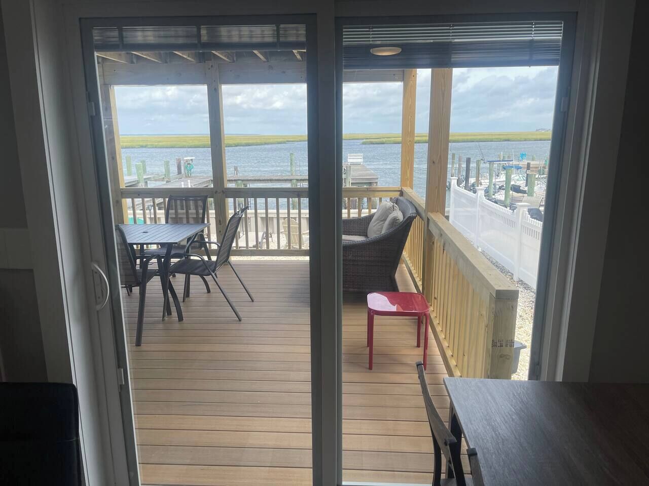 Vista desde una habitación con puerta corrediza de vidrio a una terraza de madera con mobiliario exterior.