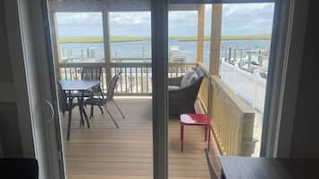 Vista desde una habitación con puerta corrediza de vidrio a una terraza de madera con mobiliario exterior.