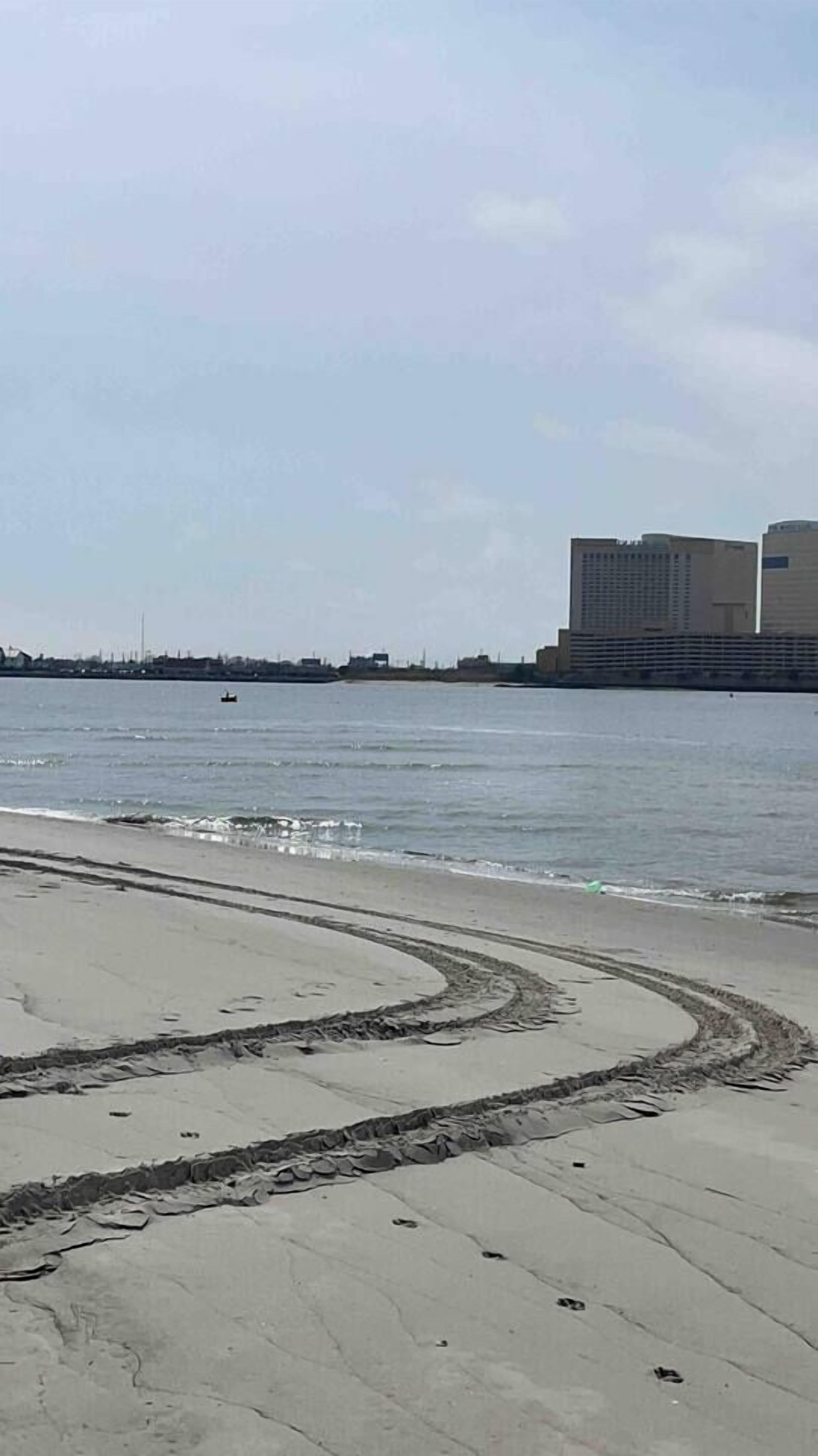 A beach with a curved sand pattern, a body of water, and distant buildings.