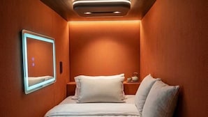 A compact hotel room with a bed, bedside tables, a wall-mounted light, and a small bedside table with a lamp. - Capsule CDMX (Mexico City)