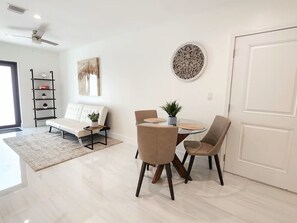 Ein modernes Wohnzimmer mit einem weißen Sofa, einem Glastisch mit vier Stühlen, einer Wanddekoration und einem Deckenventilator.