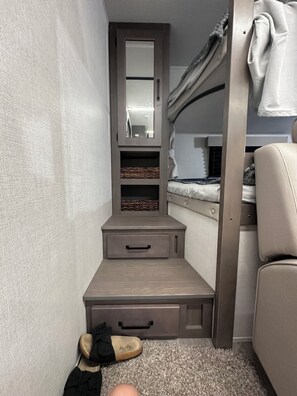 Quarto compacto com uma cama de solteiro e beliche, uma cômoda pequena com gavetas, um espelho e uma prateleira com cestos.