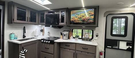 A compact kitchen with a microwave, stove, and sink, a flat-screen TV, and a window with a view of trees.