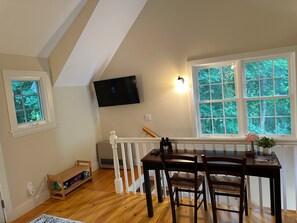 Ein Essbereich mit Holztisch und Stühlen, einem an der Wand befestigten Fernseher und einem Fenster mit Blick auf Bäume.