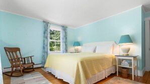 A bedroom with a bed, two bedside lamps, a rocking chair, a window with curtains, and a patterned rug. - Pom Cottage - 3BR Waterfront Cottage Near Acadia (Prospect Harbor)