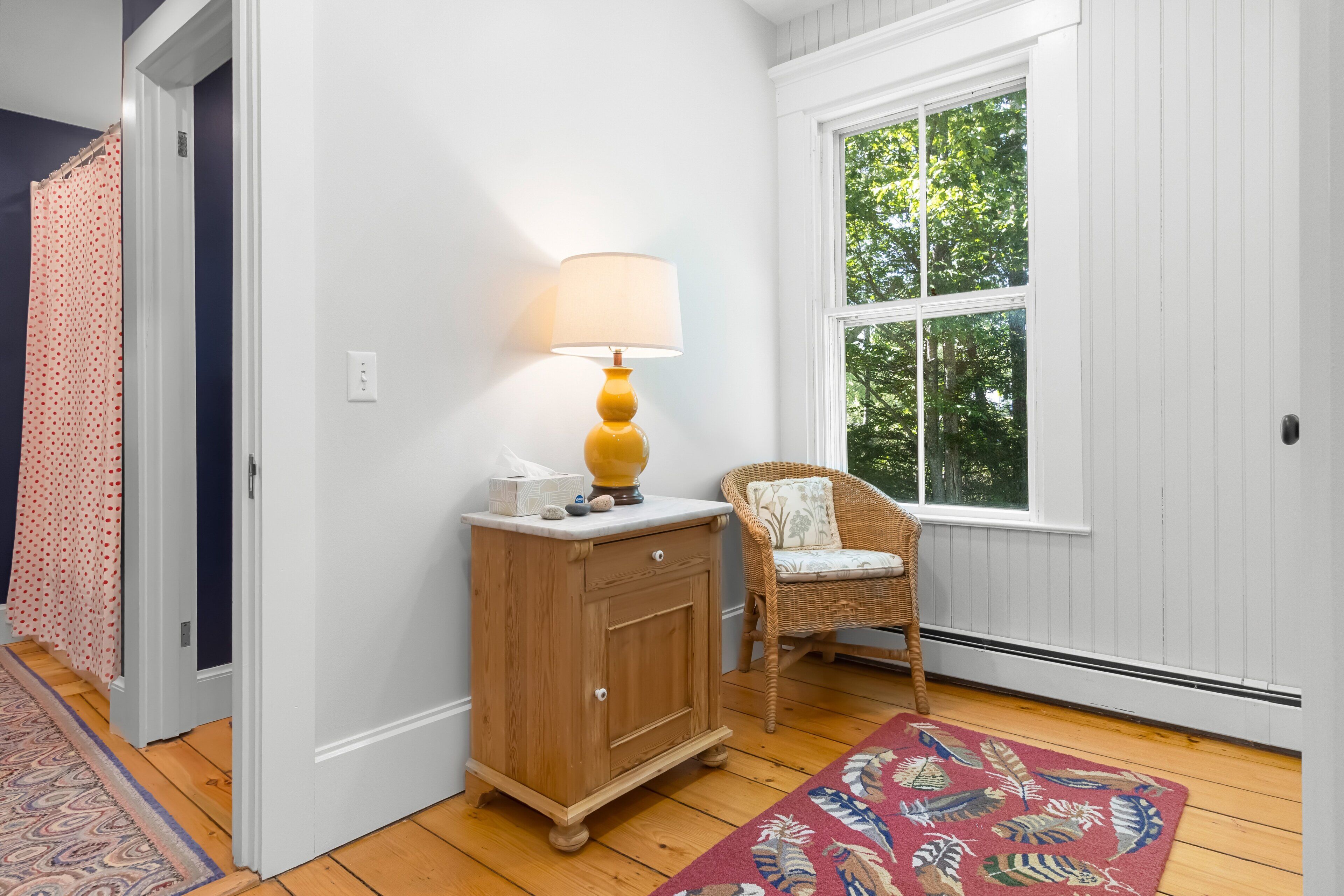 A room with a wooden cabinet, a wicker chair, a patterned rug, and a window with a view of trees.