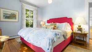 A bedroom with a red headboard, a large bed with patterned bedding, a wicker chair, a nightstand with a lamp, and a window with curtains. - Pom Cottage - 3BR Waterfront Cottage Near Acadia (Prospect Harbor)