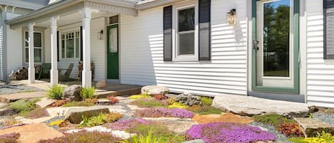 A white house with a green door, a porch with chairs, and a garden with various plants and rocks.