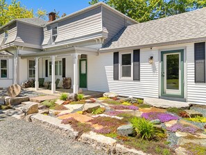 Ein zweigeschossiges Haus mit einer Veranda, einer grünen Tür und einem Garten mit verschiedenen Pflanzen und Steinen.