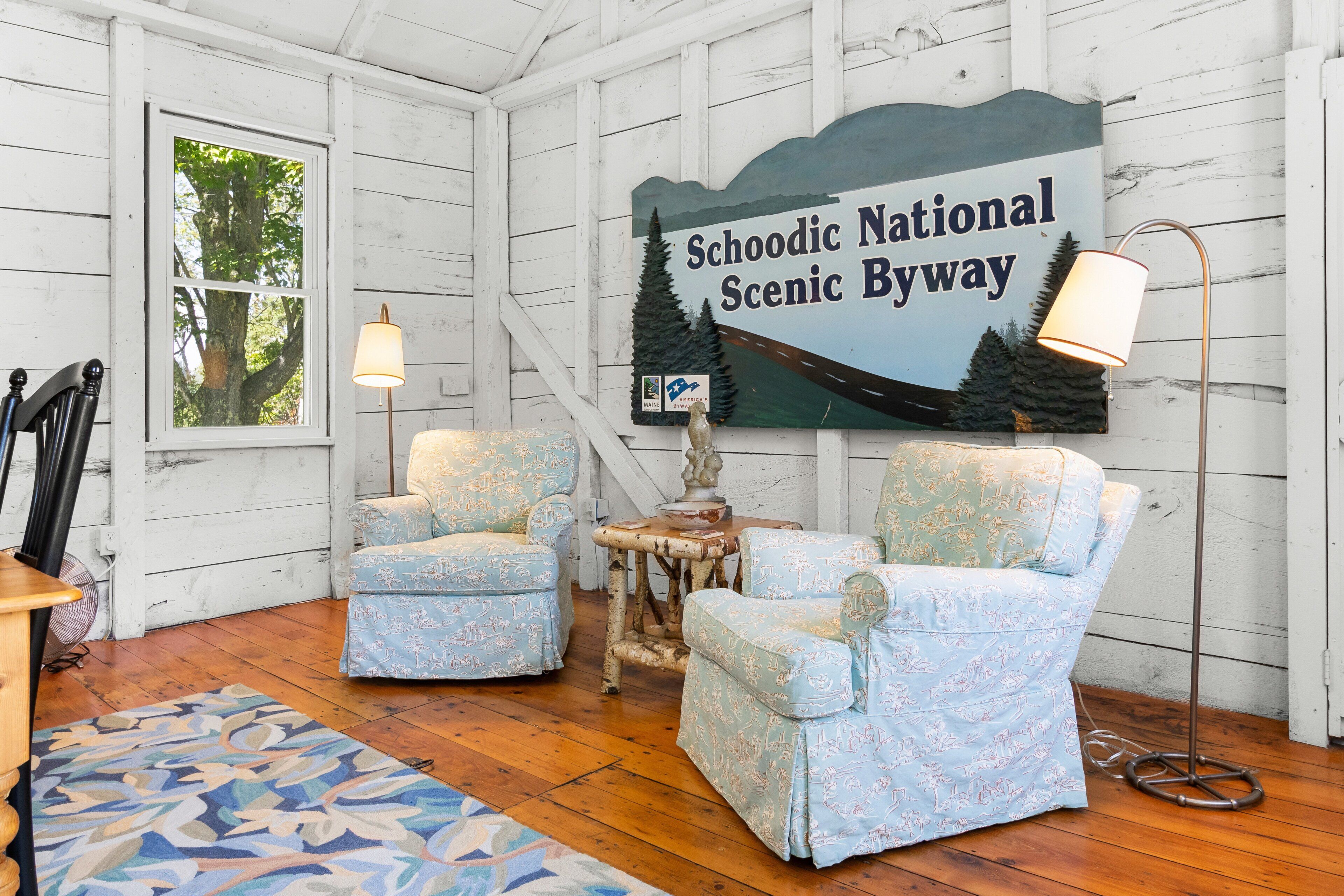Ein rustikaler Raum mit Holzwänden, zwei gemusterten Sesseln, einem kleinen Tisch, einer Lampe und einem Schild mit der Aufschrift 'Schoodic National Scenic Byway'.