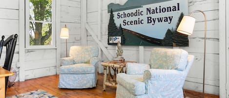 Um quarto rústico com paredes de madeira, dois poltronas estampadas, uma mesa pequena, um abajur e uma placa com a inscrição 'Schoodic National Scenic Byway'.