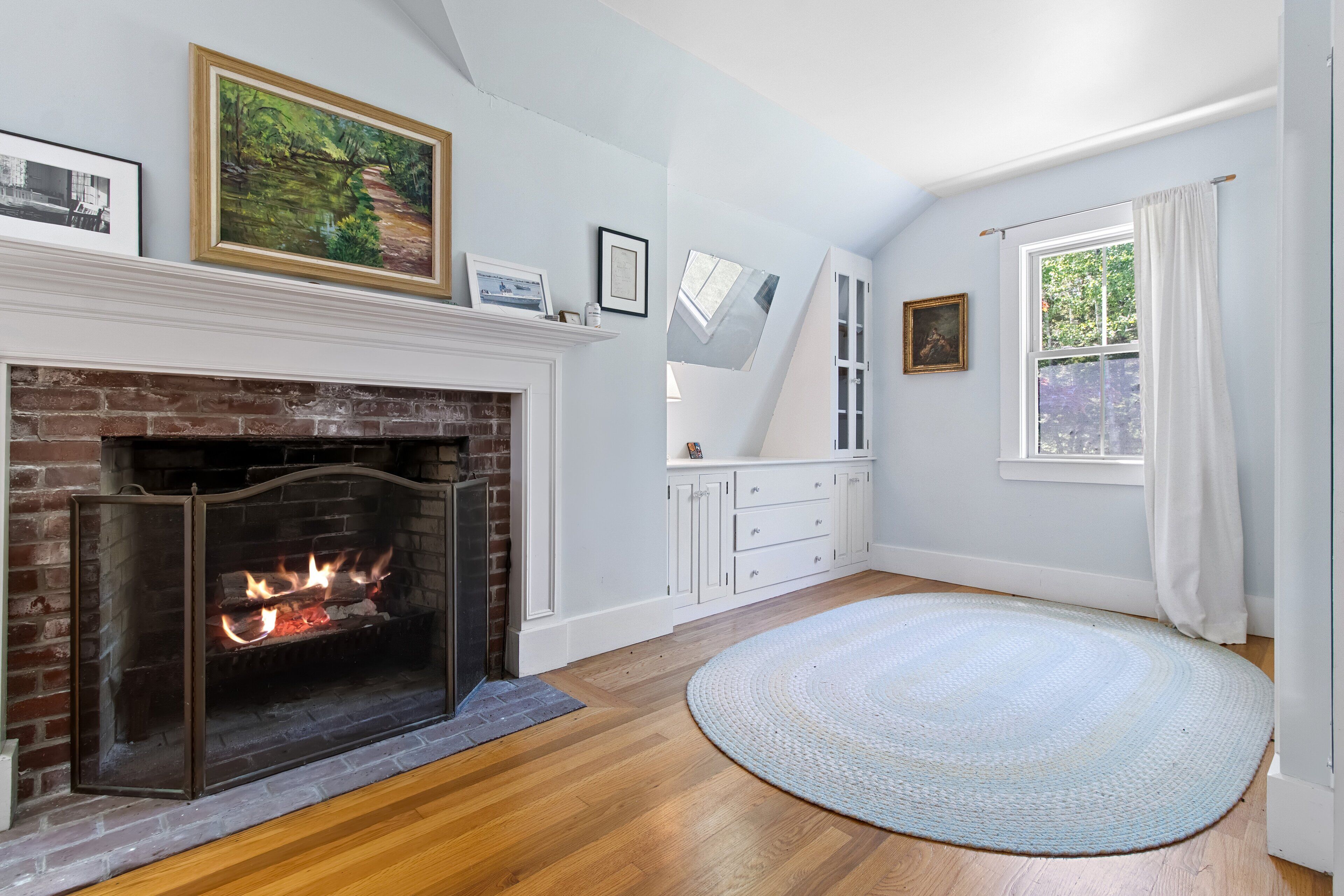 A room with a fireplace, wooden flooring, a round rug, and a window with curtains.