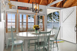 A dining area with a round table, four chairs, a vase with sunflowers, and a sign reading 'The Mermaid Lounge.' - Sunset Cottage: Bayfront 3BR Cottage With Beach Access, Fire Pit, Wifi, and Acadia Nearby (Trenton)