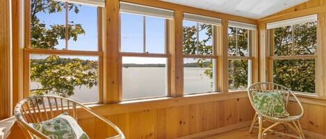 A room with wooden walls and large windows, two wicker chairs with cushions, and a view of trees and water outside.