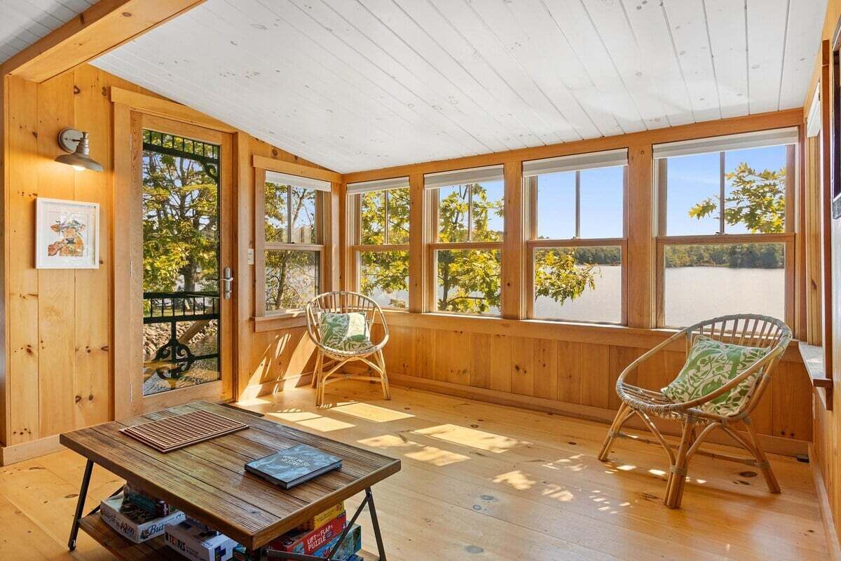 A wooden-paneled room with a table, two chairs, and large windows offering a view of trees and a body of water.