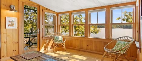 A wooden-paneled room with a table, two chairs, and large windows offering a view of trees and a body of water.