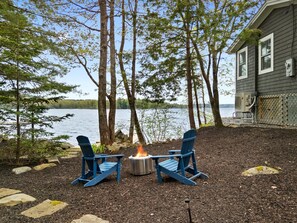 A lakeside deck with two Adirondack chairs, a fire pit, and a house in the background. - 2BR Maine Camp w/ Dock, Lake Access & AC (Nobleboro)