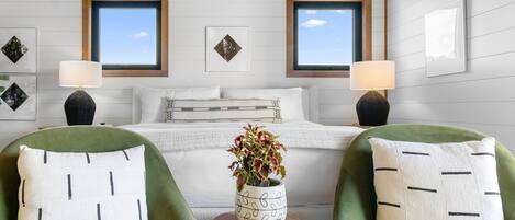 A bedroom with a bed, two chairs, a small table, and two windows.