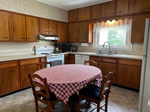 A kitchen with wooden cabinets, a stove, a dishwasher, and a window with a lace curtain. - One Atlantic 5BR Harbor View Maine Home, Close to Town, Deck (Boothbay Harbor)