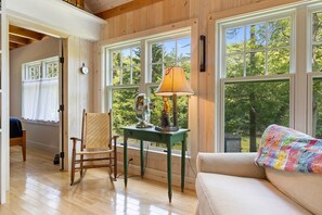 A room with a wooden floor, a rocking chair, a table with a lamp, a sofa, and large windows with a view of trees. - The Boathouse (Bath)