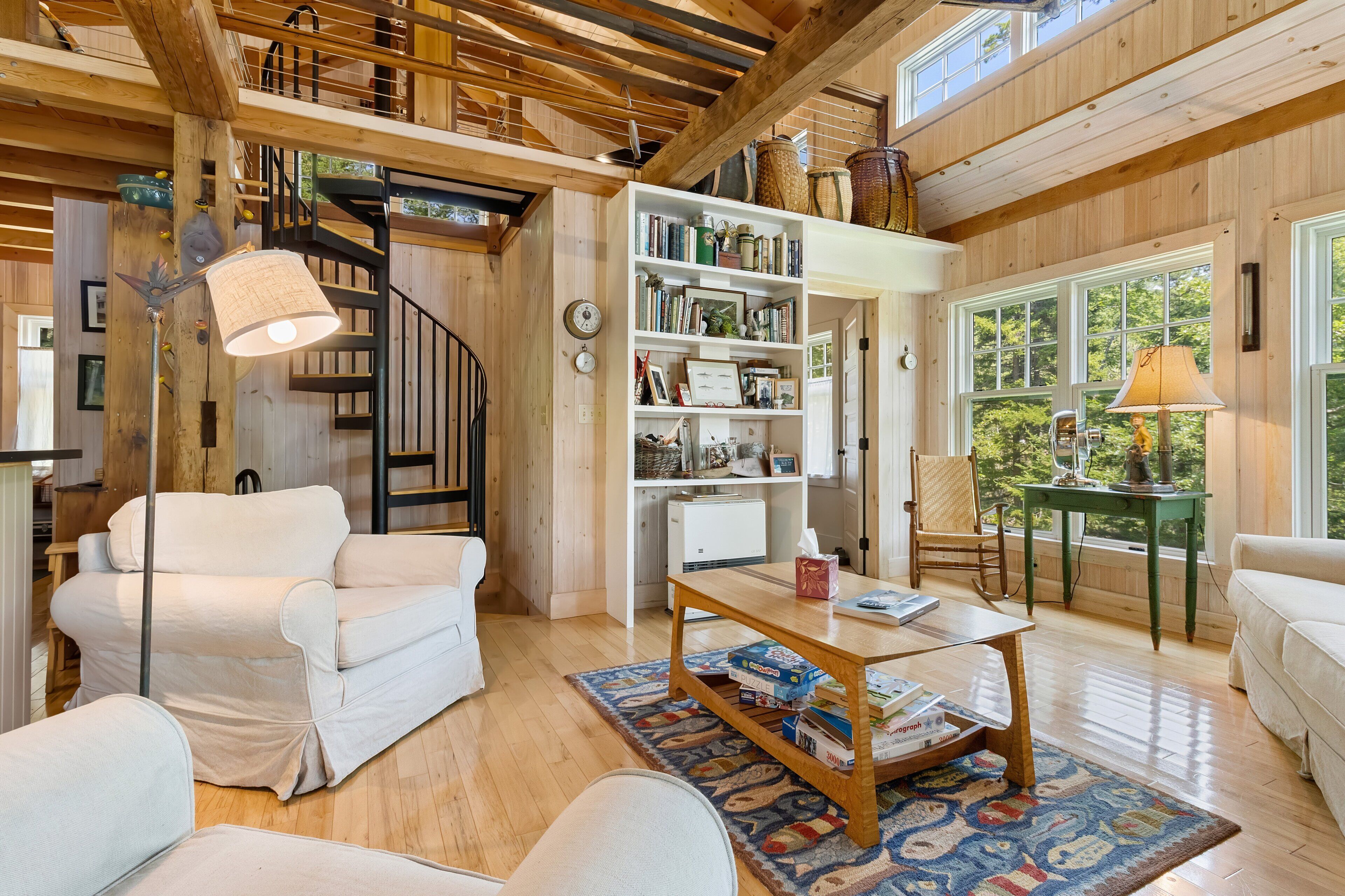 A spacious living area with a wooden ceiling, a bookshelf, a white sofa, a coffee table, and a view through large windows.