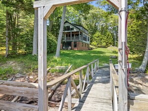 A wooden bridge leading to a two-story house with a porch, surrounded by a forest. - The Boathouse (Bath)