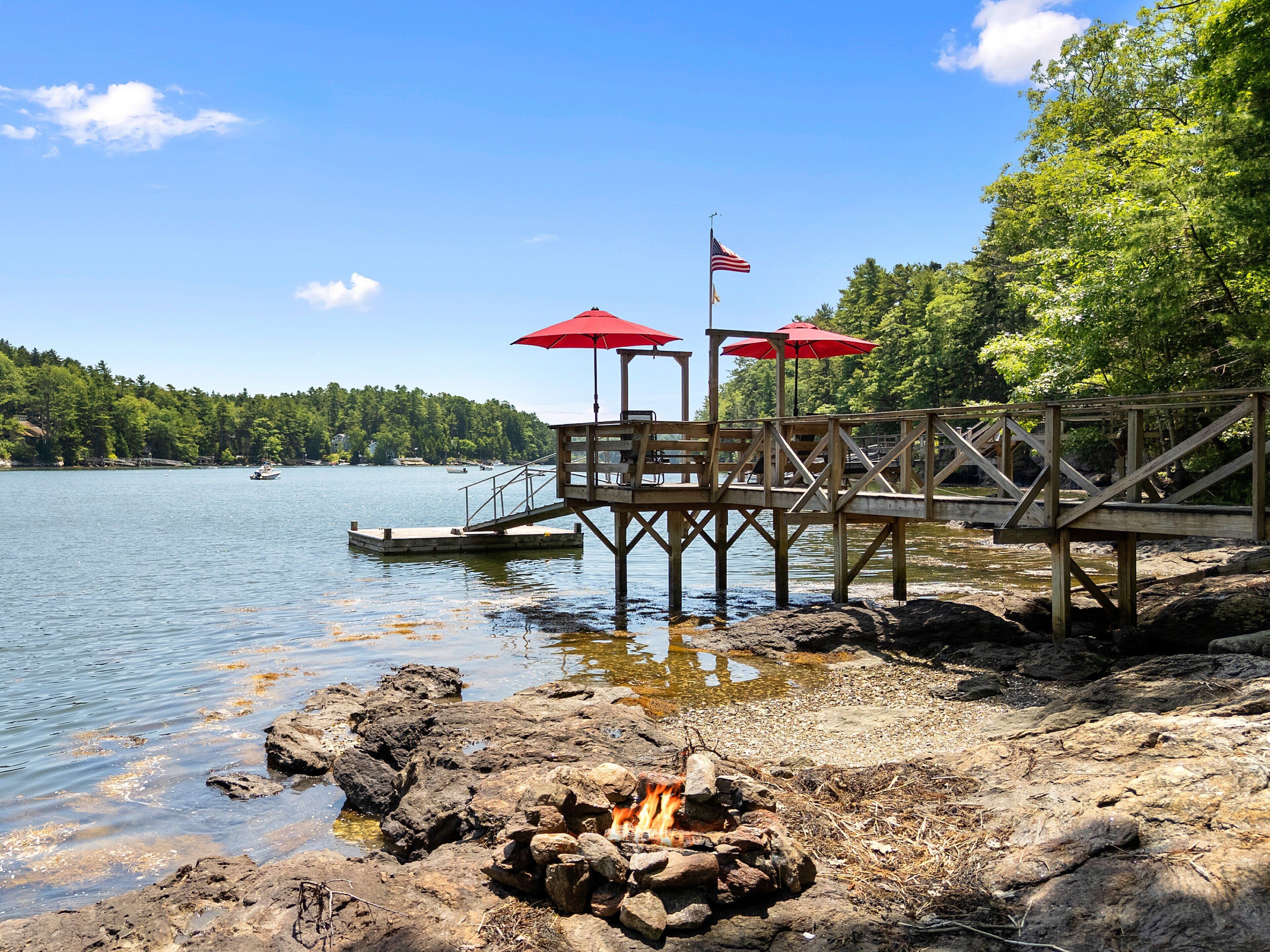A wooden dock with a fire pit and red umbrellas, overlooking a calm lake with boats and a forested shoreline.