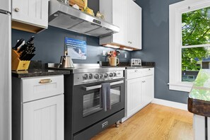 A kitchen with a stainless steel oven, white cabinets, a window with a view of trees, and a sign on the wall. - La Maisonette - Historic Renovated 6 BR Downtown Bar Harbor (Bar Harbor)