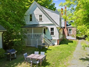 Une maison à deux étages avec un porche, un coin repas aménagé avec du mobilier d’extérieur et un chemin menant à la maison.