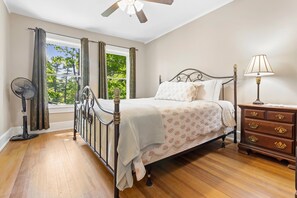 Ein Schlafzimmer mit einem Bett, einem Nachttisch, einer Lampe, einem Ventilator und einem Fenster mit Vorhängen.