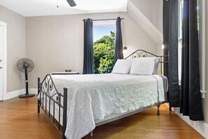 Ein Schlafzimmer mit einem Bett, einem Standventilator und einem Fenster mit Vorhängen.