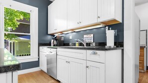 A kitchen with white cabinets, a black countertop, and a window with a view of a house and trees. - La Maisonette - Historic Renovated 6 BR Downtown Bar Harbor (Bar Harbor)