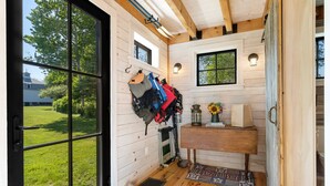 A wooden cabin interior with a large window, a wall-mounted luggage rack, a small wooden table with a vase, and a blue mat at the entrance. - Aril Cottage-waterfront Escape With Ocean Views (Prospect Harbor)