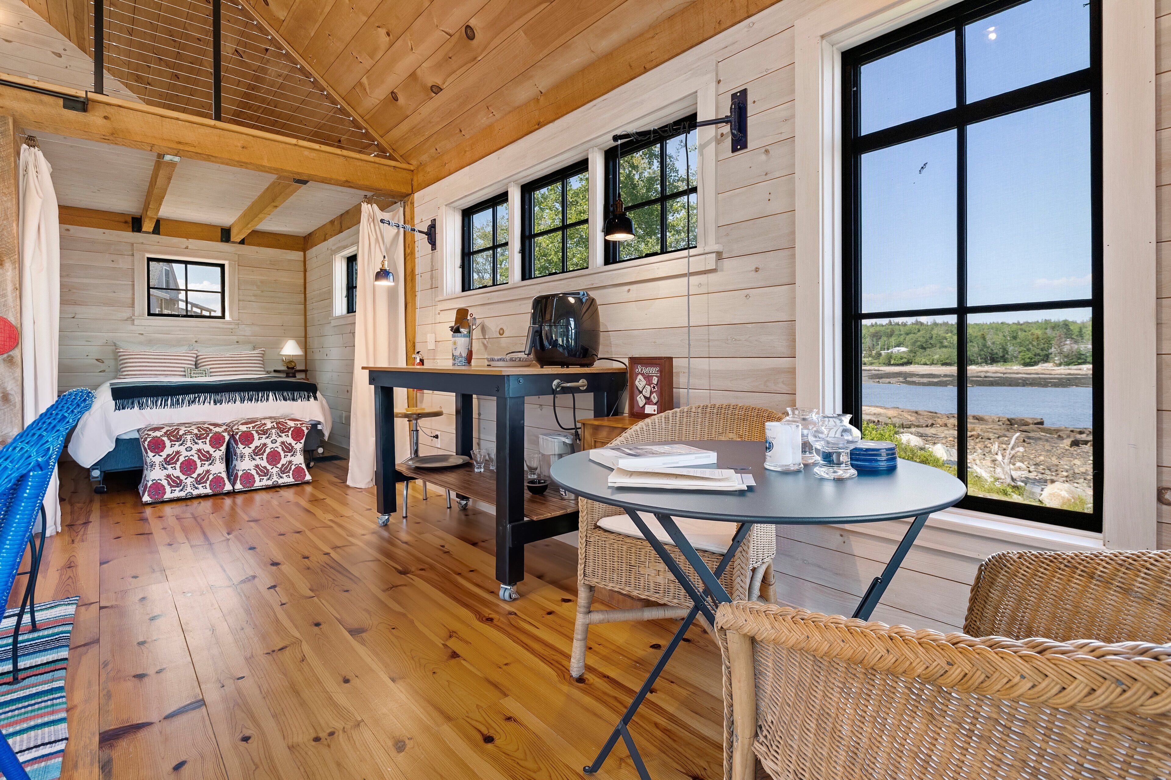 A cozy cabin interior with a wooden floor, a dining table, a sofa, and a view of a lake.
