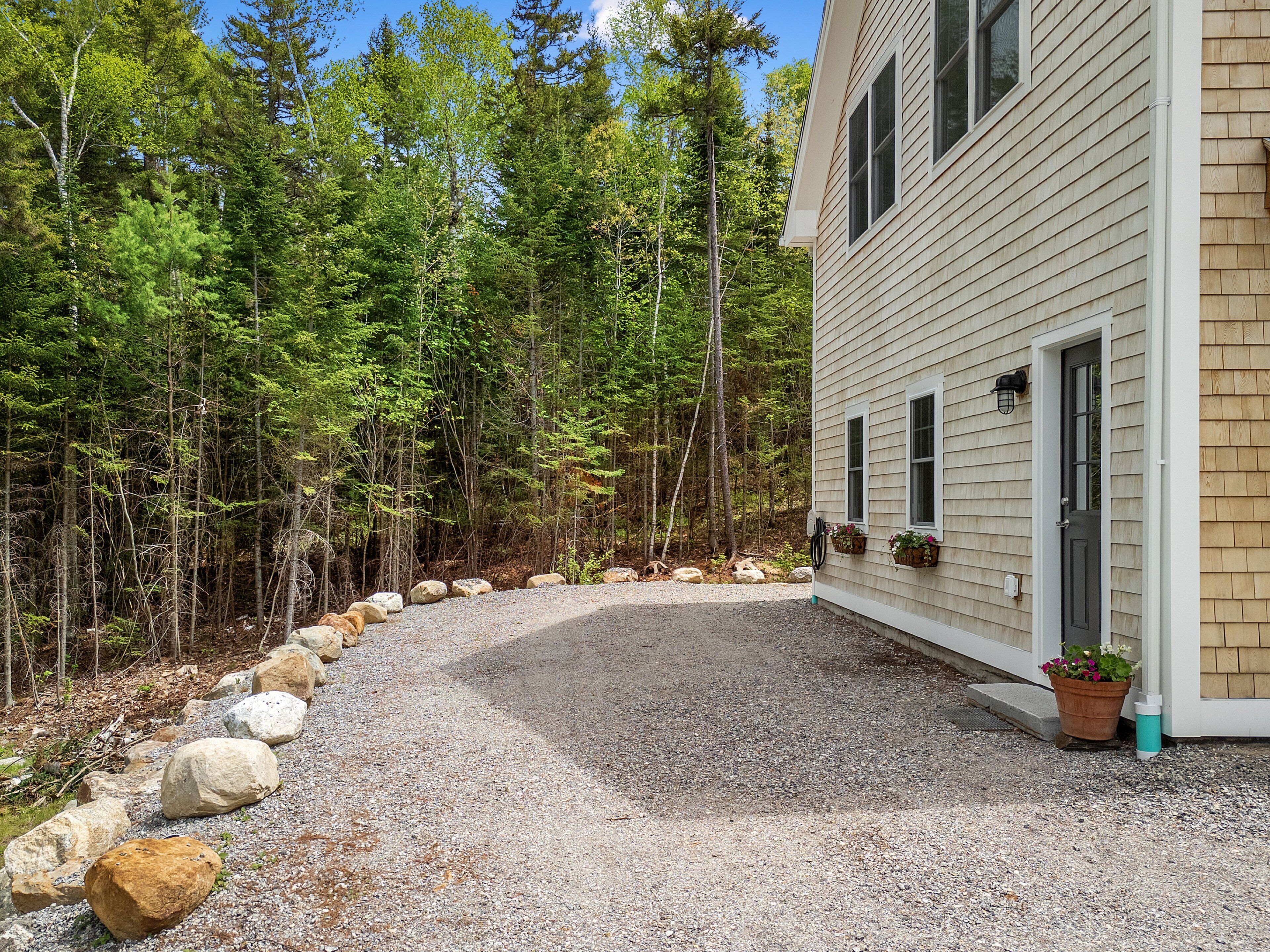 A gravel driveway leading to a house with a wooden exterior, surrounded by a forested area with rocks along the path.