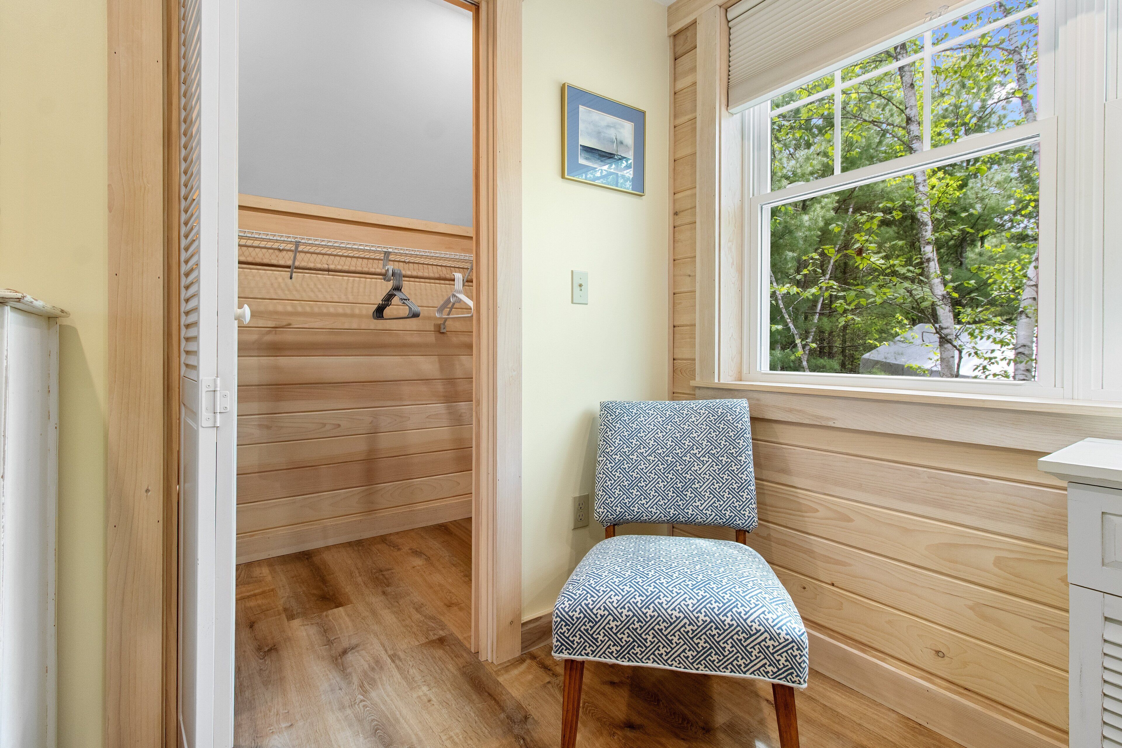 A room with a wooden floor, a chair with a patterned cushion, a window with a view of trees, and a door leading to a closet with hanging clothes.
