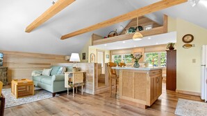 A spacious loft with a high ceiling, wooden beams, a kitchen area with a central island, a dining area with a sofa and chairs, and a large window providing natural light. - Riverbend Perch - River-view Retreat for 2 With Kitchen, Patio, AC, Wi-fi (Cushing)