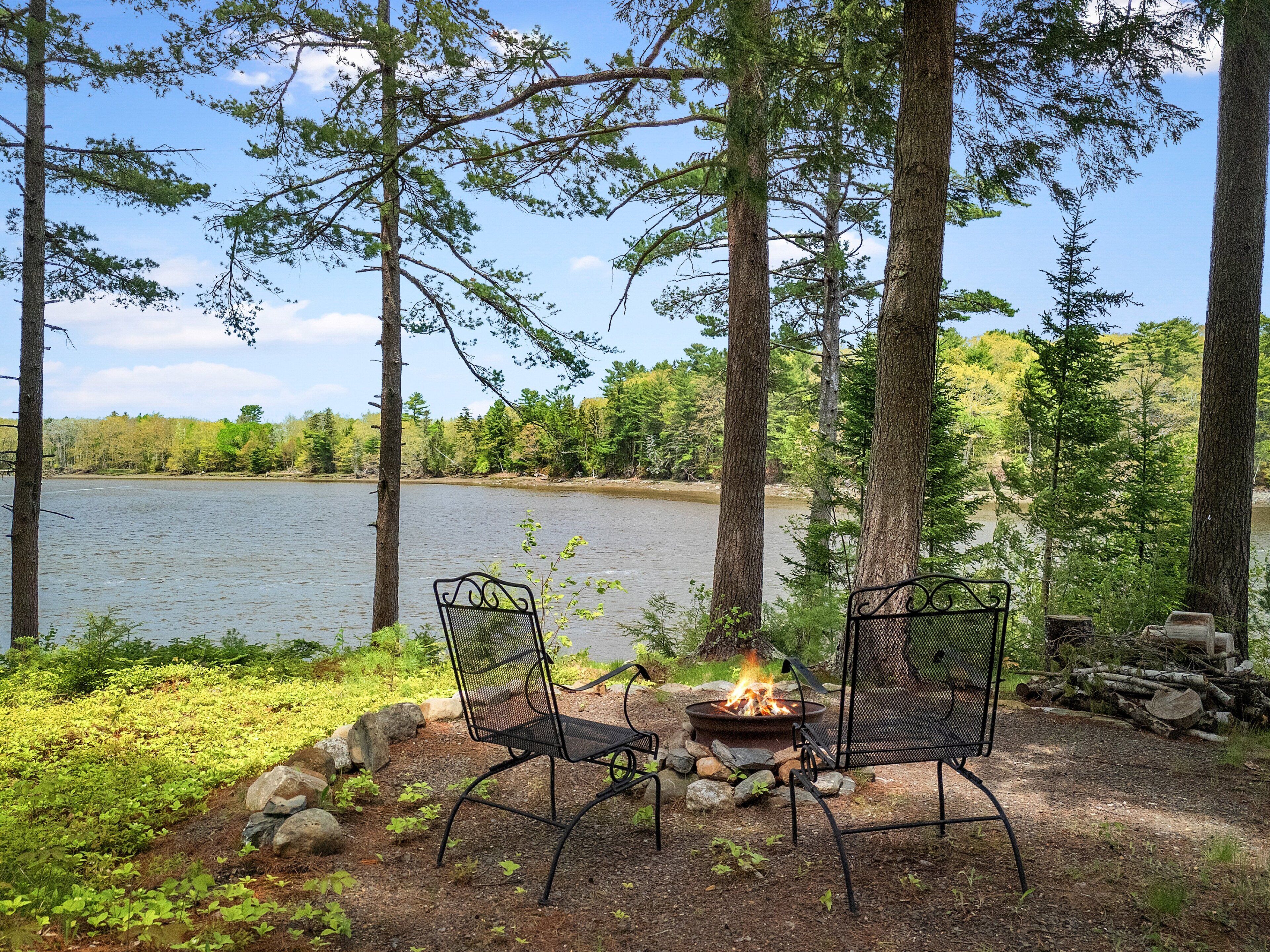 A lakeside outdoor seating area with two chairs and a fire pit.