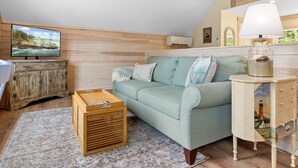 A cozy living room with a wooden floor, a sofa, a wooden cabinet, a television, and a lamp. - Riverbend Perch - River-view Retreat for 2 With Kitchen, Patio, AC, Wi-fi (Cushing)