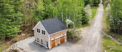 A two-story house with a gravel driveway and a stone wall, surrounded by dense forest.