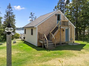 A small wooden cabin with a staircase leading to a deck, situated near a body of water with trees in the background. - Capstone Cottage - Quintessential Cabin on Bay (Prospect Harbor)