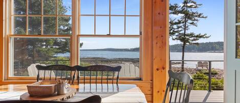 A wooden interior with a table and chairs, large windows with a view of a beach and trees.