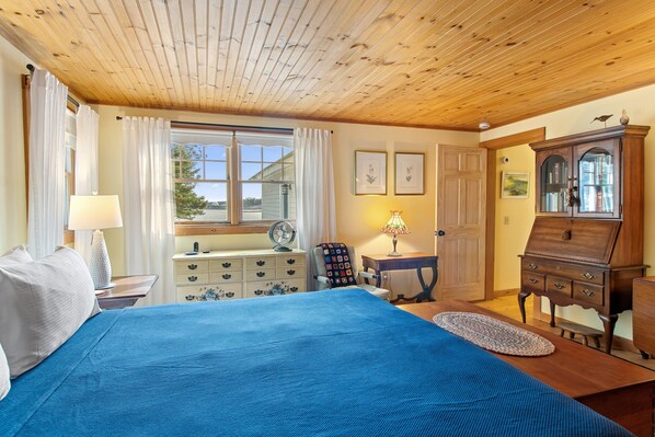 Una habitación con techo de madera, una cama con colcha azul, un aparador, una silla y una ventana con cortinas.