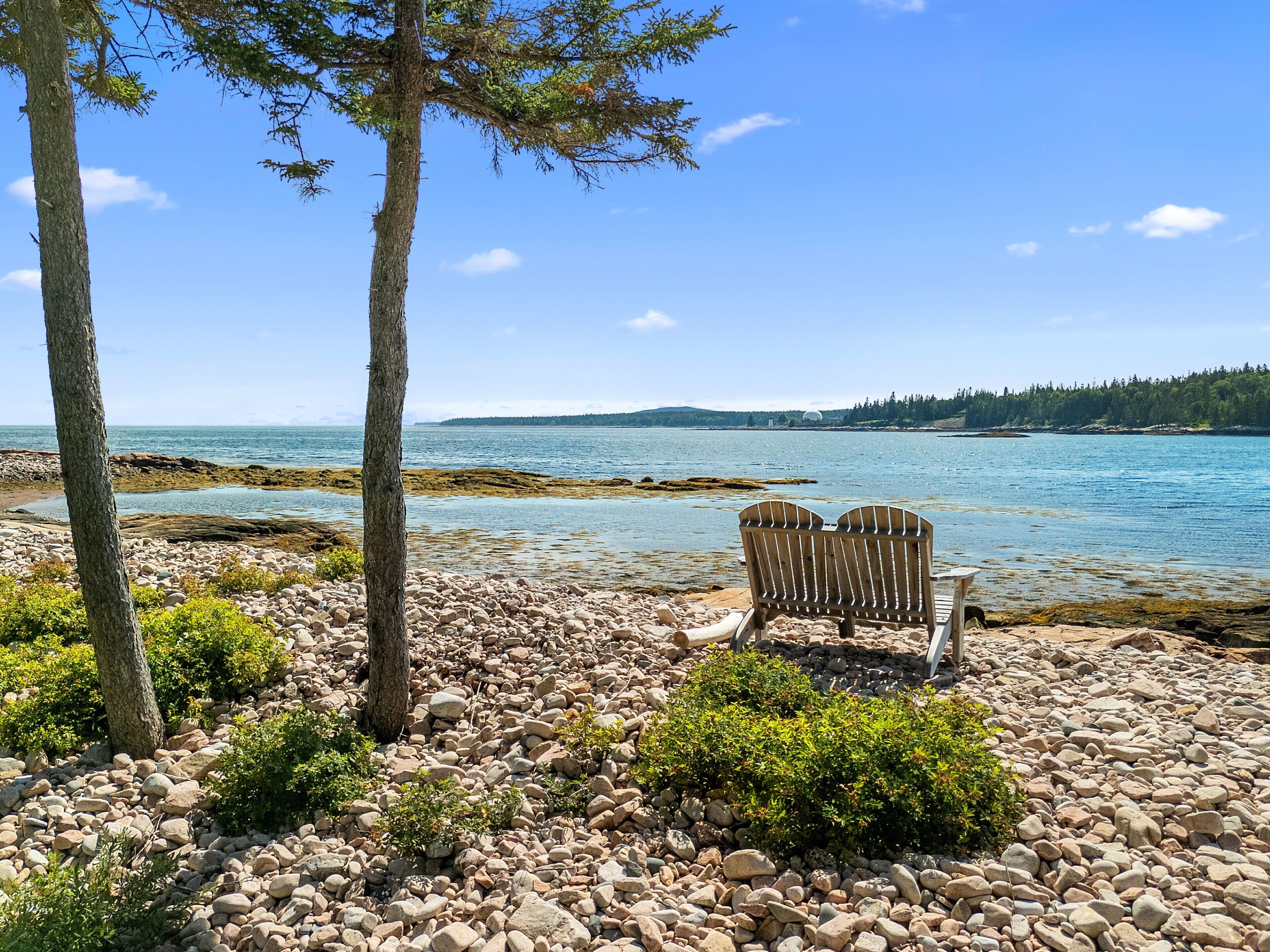A rocky beach with two wooden chairs facing the sea, trees, and a clear sky.