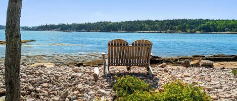 A wooden bench on a rocky shore with a view of a calm lake and forested land in the distance.