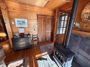 Interior de una cabaña de madera con chimenea, cómoda, silla y puerta con ventana.