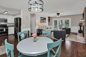 Una cocina moderna con electrodomésticos de acero inoxidable, un comedor con una mesa redonda y sillas color turquesa, y una sala de estar con sofá y chimenea.